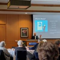 A man speaks to a crowd in front of a projector that says, "What Is Media?"